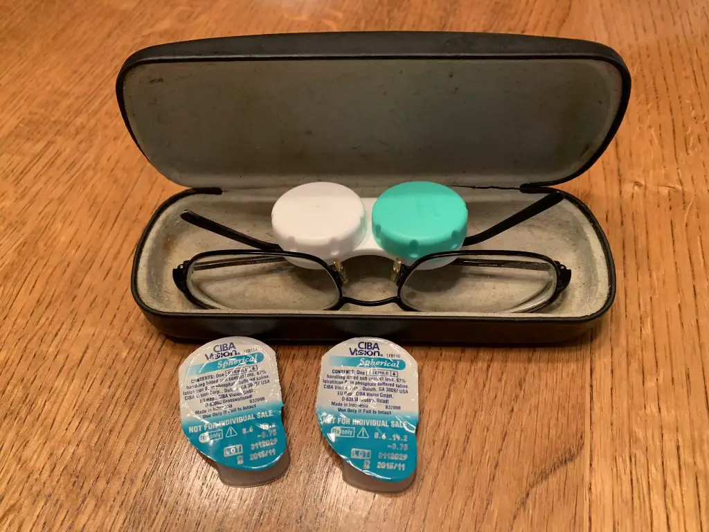 glasses and a contact case sitting in a glass case, with extra contact packets sitting in front of them